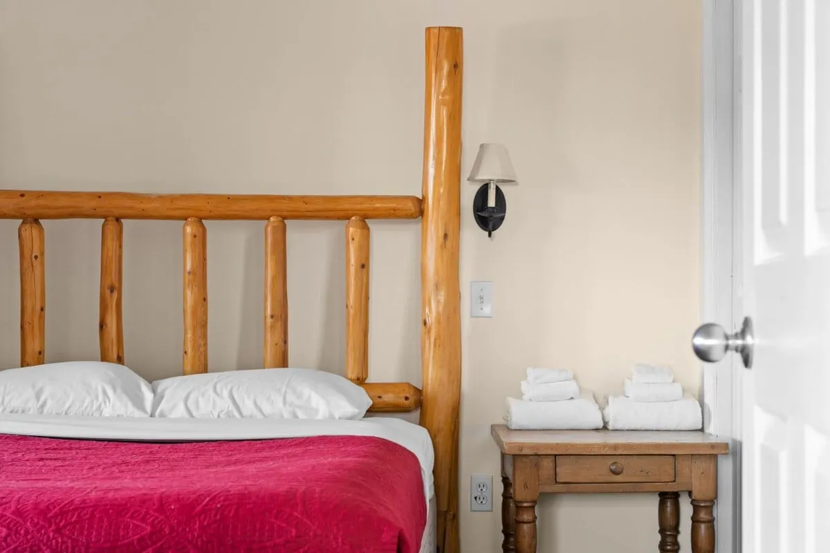 A cozy bedroom with a wooden bed frame, topped with a rich red bedspread. A nightstand with neatly folded towels hints at thoughtful hospitality. Soft natural light streams in, creating a tranquil atmosphere ideal for relaxation or morning routines.