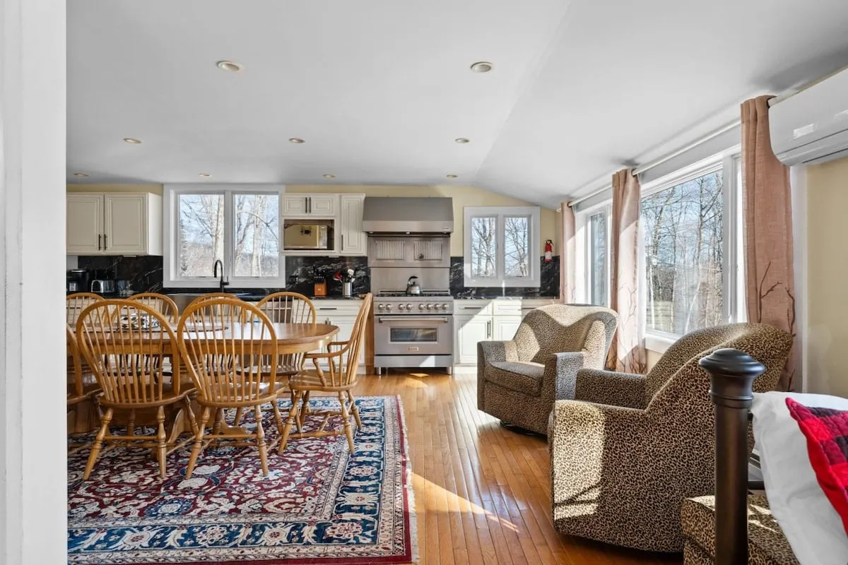 A warm, inviting dining space connected to a modern kitchen and comfortable seating area. The large dining table sits beside sizable windows, allowing for plenty of natural light and views of the outdoors. The arrangement promotes group meals and easy socialization.
