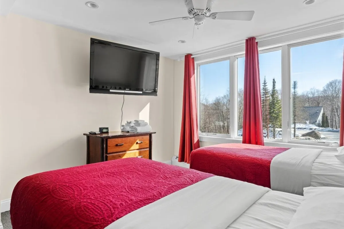 This image features a cozy bedroom with two red beds positioned near large windows draped with curtains. The windows offer a view outside, inviting in natural light and connecting the interior to the outdoors. A TV on the wall and a wooden dresser complete the comfortable setting, ideal for relaxation after a day of exploring.