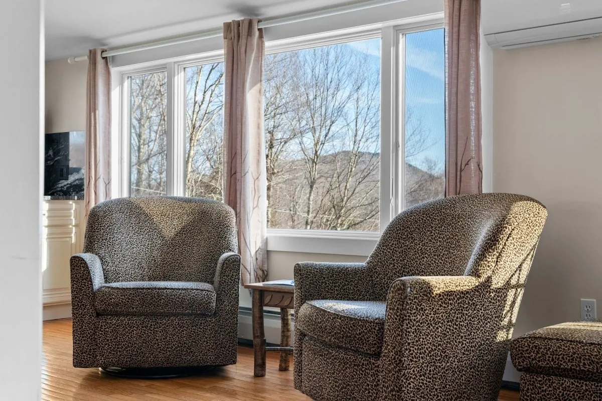A relaxed sitting area with leopard print chairs, a wooden side table, and expansive windows offering scenic mountain views. The arrangement invites quiet moments of reading or casual conversations while appreciating the outdoors from within.