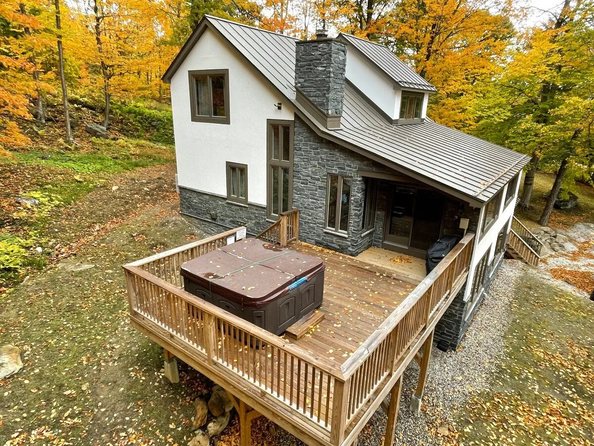 An exterior perspective featuring the house’s modern architecture, the spacious wooden deck, and the outdoor hot tub. The image is set against brilliant autumn foliage, conveying the deck’s connection to the wilderness and offering a sense of the property’s outdoor entertainment and relaxation possibilities.