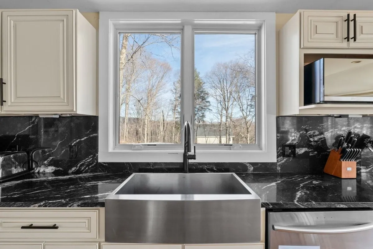 A modern, fully equipped kitchen with black marble countertops, a stainless steel sink, and large windows. The layout is designed for both meal preparation and socializing, offering ample space for group cooking and entertaining.