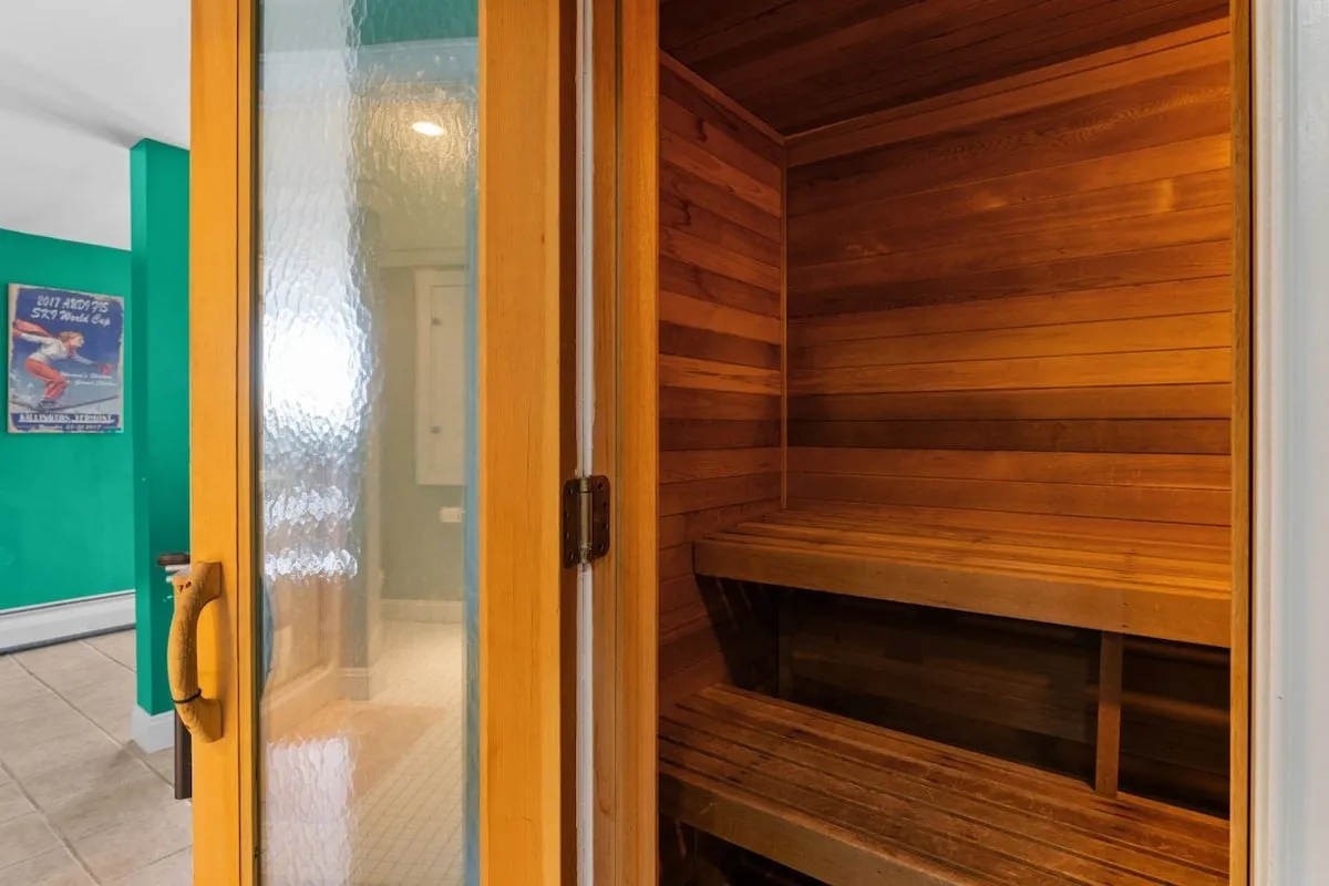 The image features the inviting interior of a wooden cedar sauna, with warm wood tones, a glass door, and subtle lighting that underscores the space’s calming atmosphere. The bench seating highlights space for multiple guests, suggesting social relaxation or personal wellness routines.