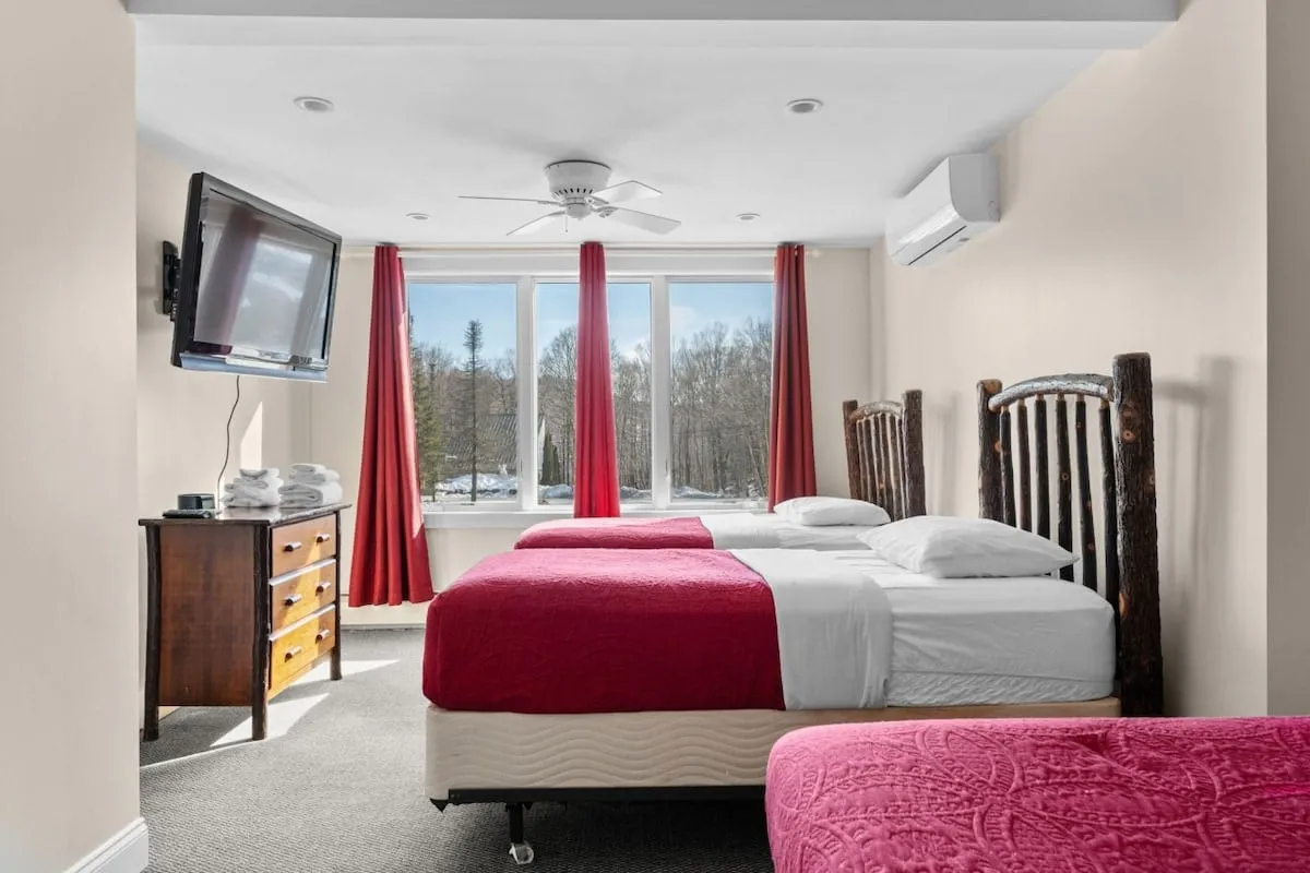 This room features two beds adorned with vibrant red bedspreads and sits beside large windows with deep red curtains, allowing for ample natural light. A wooden dresser provides functional storage, making it ideal for friends or family sharing the space.