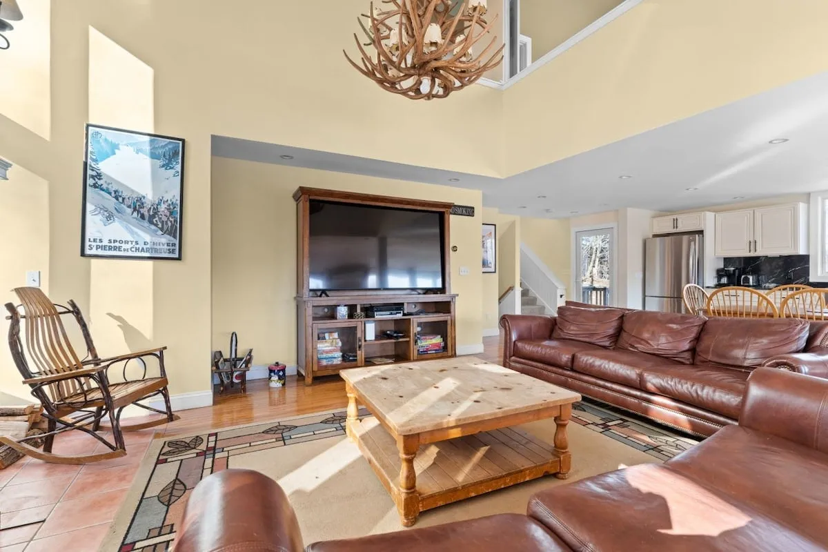 A spacious living area featuring leather sofas, a wooden coffee table, a large television, and a striking antler chandelier. The space is arranged around a cozy, oversized wood-burning fireplace (not directly visible but implied by the arrangement), perfect for social gatherings after a day on the slopes.