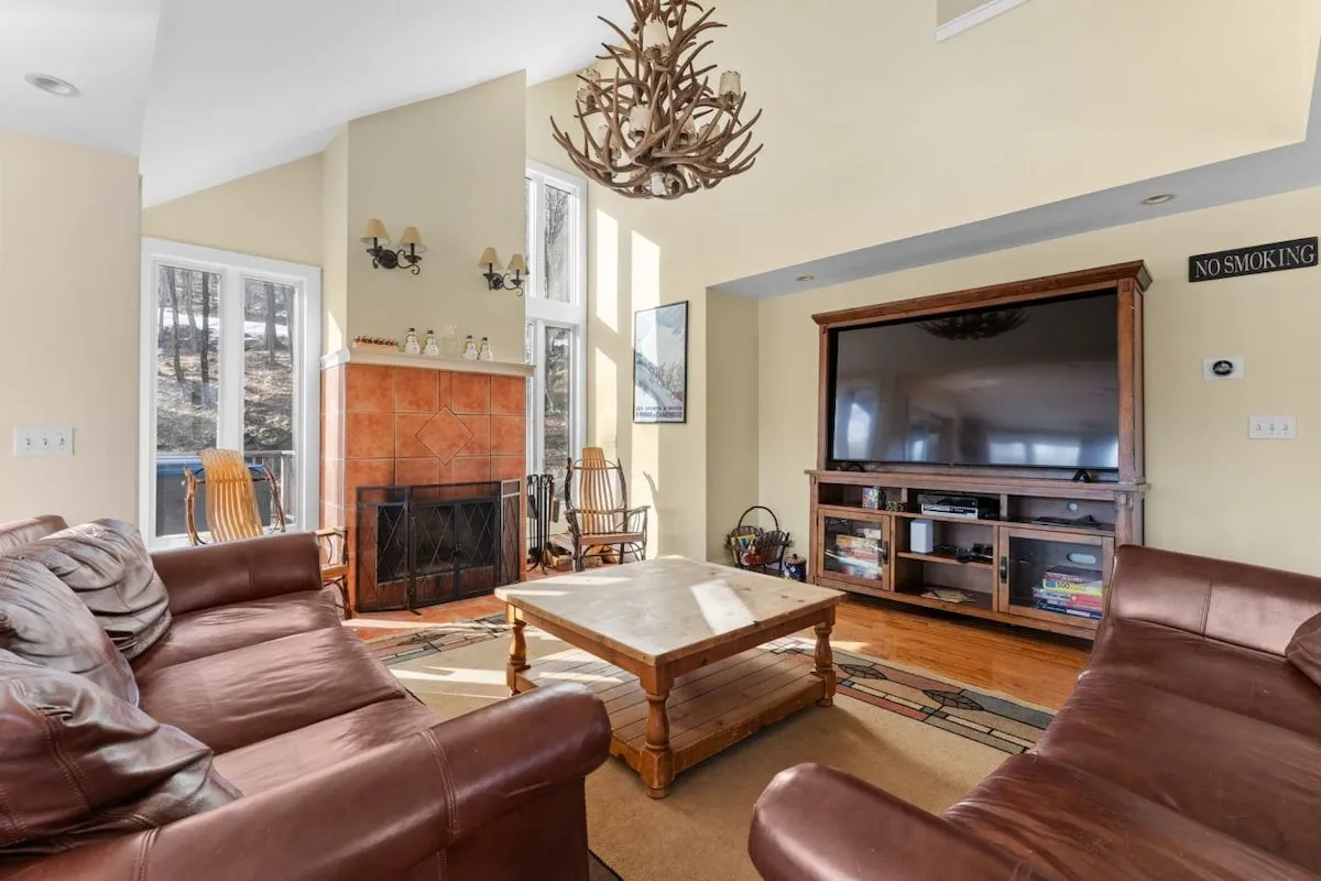 A cozy living room scene with leather sofas arranged around a stone fireplace, an antler chandelier overhead, a large smart TV, and a rustic wooden coffee table. Ample natural light enters through large windows, enhancing the welcoming atmosphere.