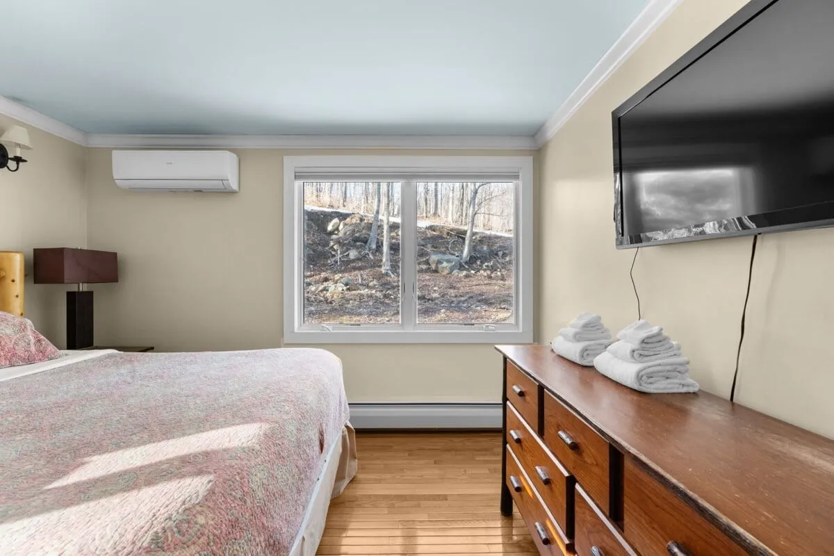 A serene bedroom with a quilted bed facing a large window. The view through the window brings in lush, natural scenery, while a wooden dresser and wall-mounted TV add functional comfort. The space feels peaceful, welcoming guests and their pets to enjoy both comfort and a close relationship with the outdoors.