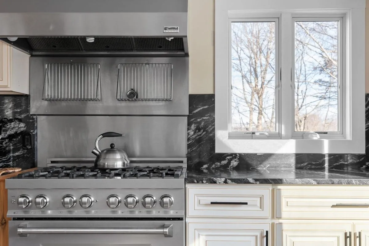 A warm and modern kitchen view centered on a stainless steel stove and black marble countertop. A window behind the stove provides a serene outlook to treetops, blending indoor comfort with a touch of nature. The countertop is ready for meal preparation and group cooking sessions.