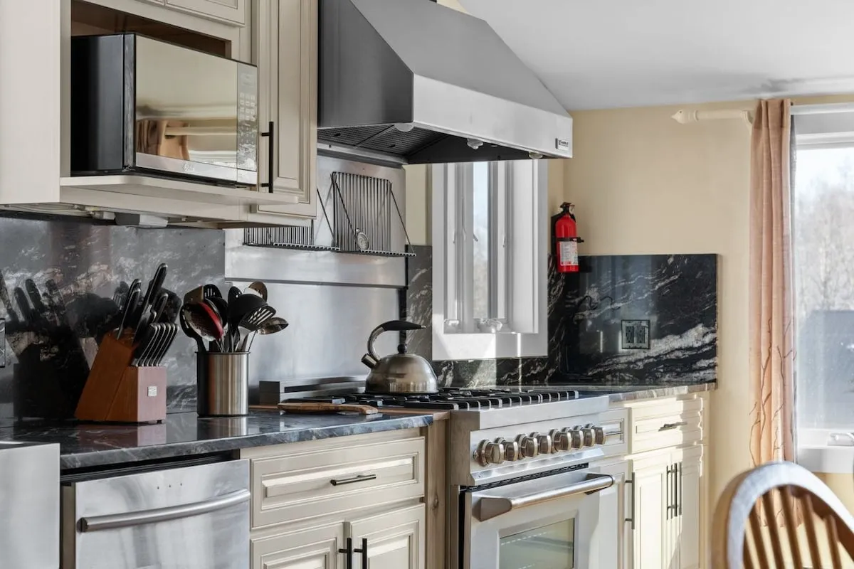 A stylish kitchen interior with a stainless steel stove, black granite countertop, and a variety of kitchen utensils visible. The scene exudes a sense of warmth and hospitality, suggesting a space designed for active cooking, entertaining, and creating family traditions.
