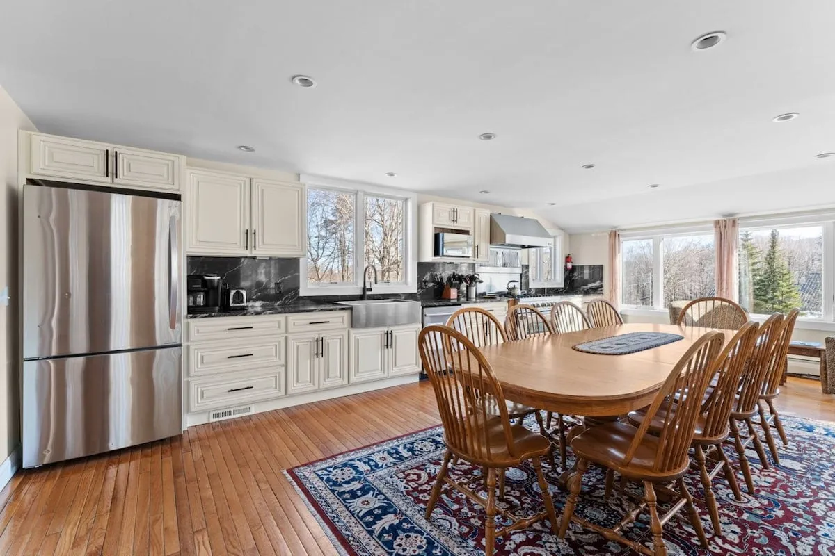 The image showcases a spacious, sunlit dining area with a large table perfect for group gatherings, an elegant kitchen island ideal for shared cooking experiences, and expansive windows framing scenic mountain views.