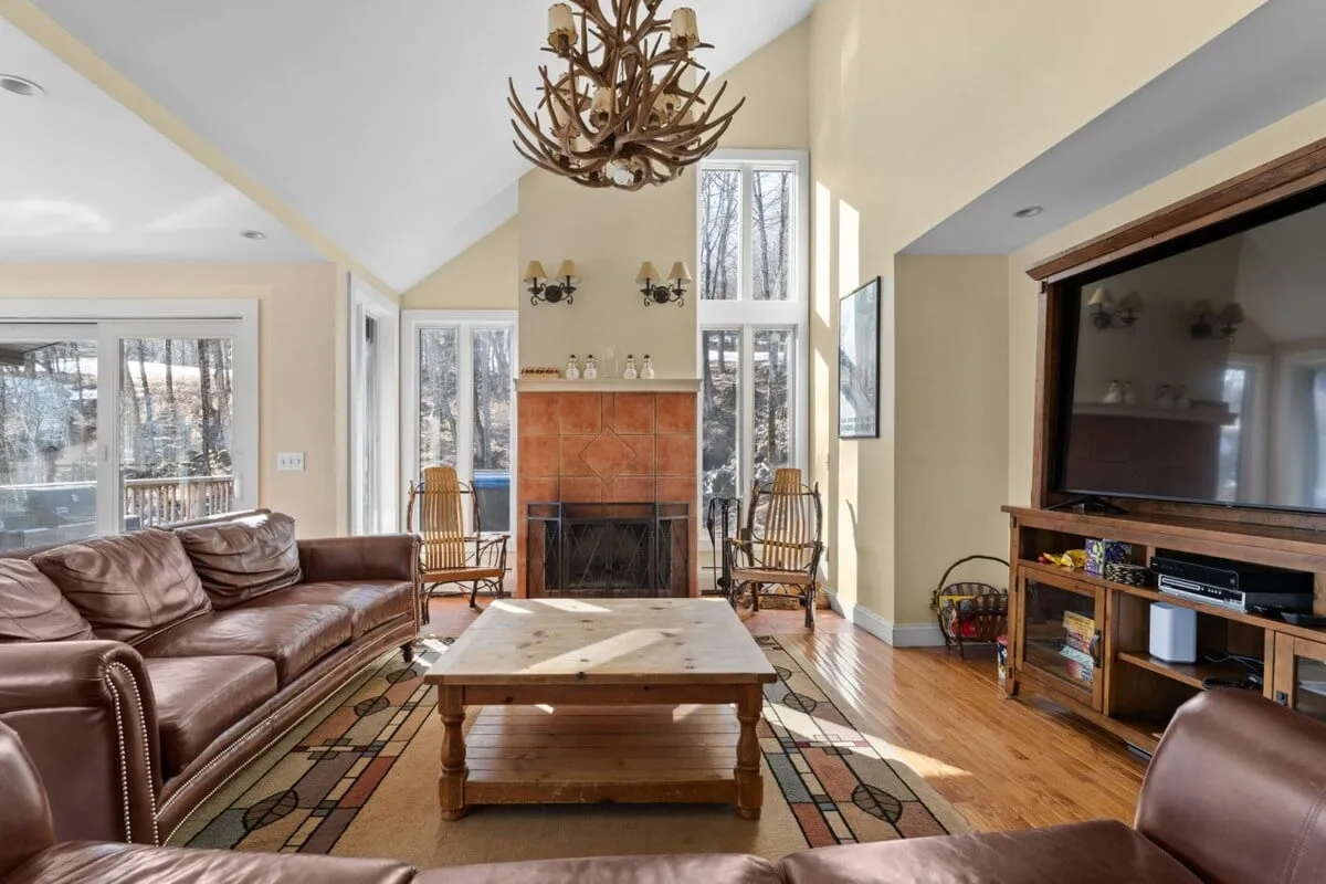 A warm, inviting living room featuring a leather sofa, antler chandelier, fireplace, and large windows that frame the outdoor view. The scene suggests relaxation and comfort after a day on the slopes.