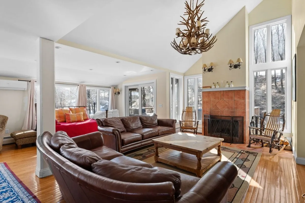 A spacious living room featuring plush leather sofas arranged around a large wood-burning fireplace. Expansive windows flood the space with natural light, while a stylish chandelier adds a touch of elegance. The room seamlessly connects to an adjacent dining area, emphasizing the open-concept design.