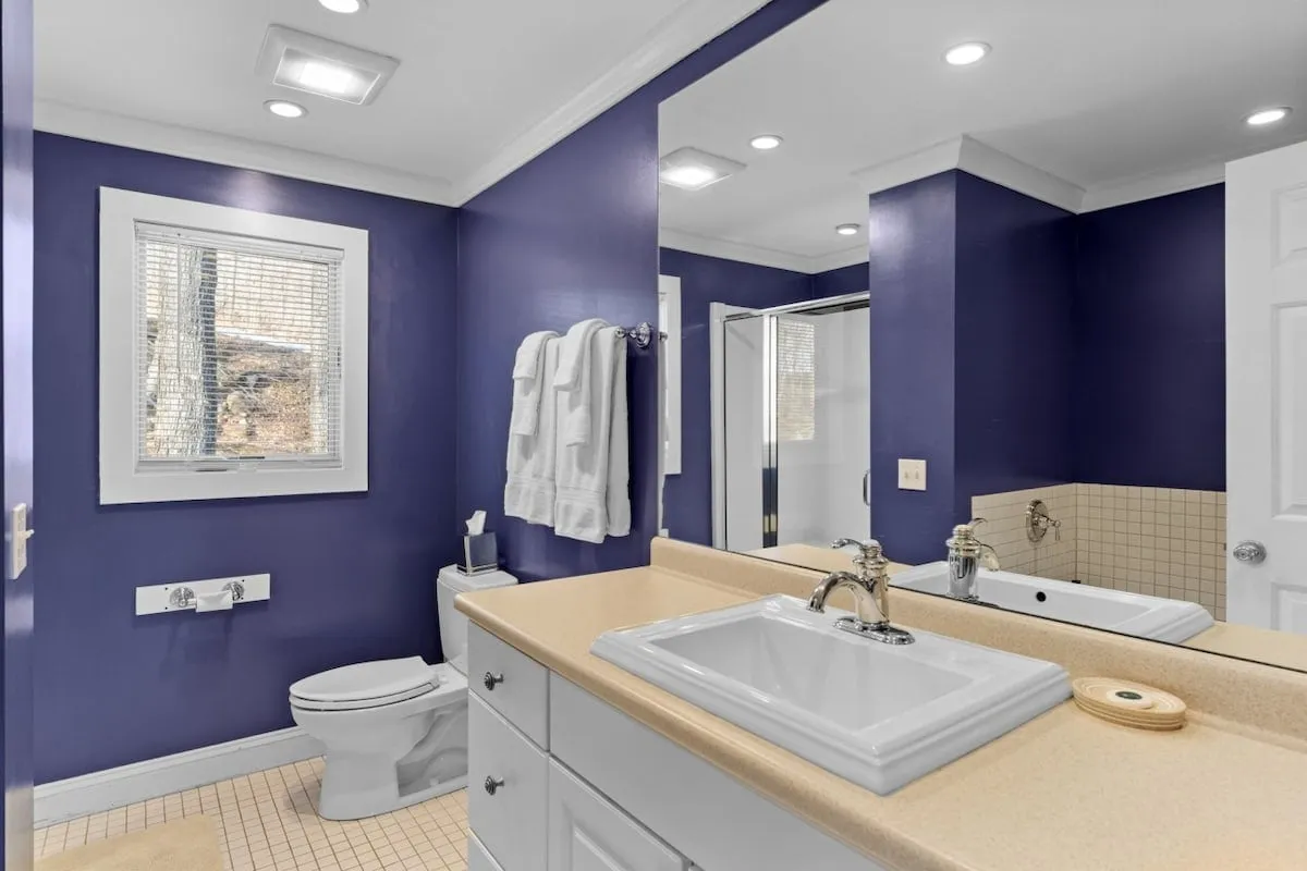 A spa-like bathroom with lavender walls, double sinks, a large mirror, modern fixtures, and a window welcoming in natural light. The ambiance is calm and refreshing, ideal for unwinding after a long day.
