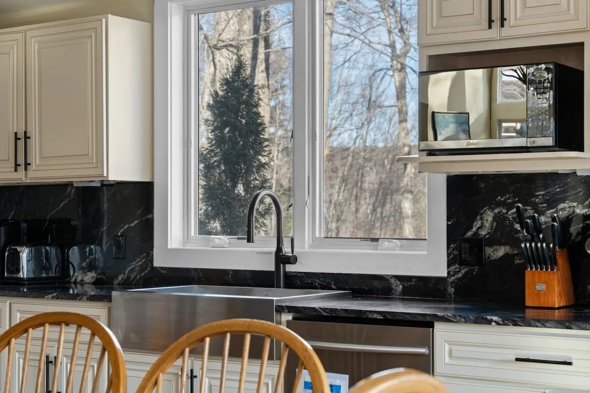 A warm and welcoming kitchen scene featuring a prominent kitchen sink beneath a window, black countertop, ample kitchen cabinets, and neatly arranged kitchen utensils. Natural light streams through the window, highlighting the cozy and functional aspects of the space. The visible layout suggests a room designed for both cooking and socializing.